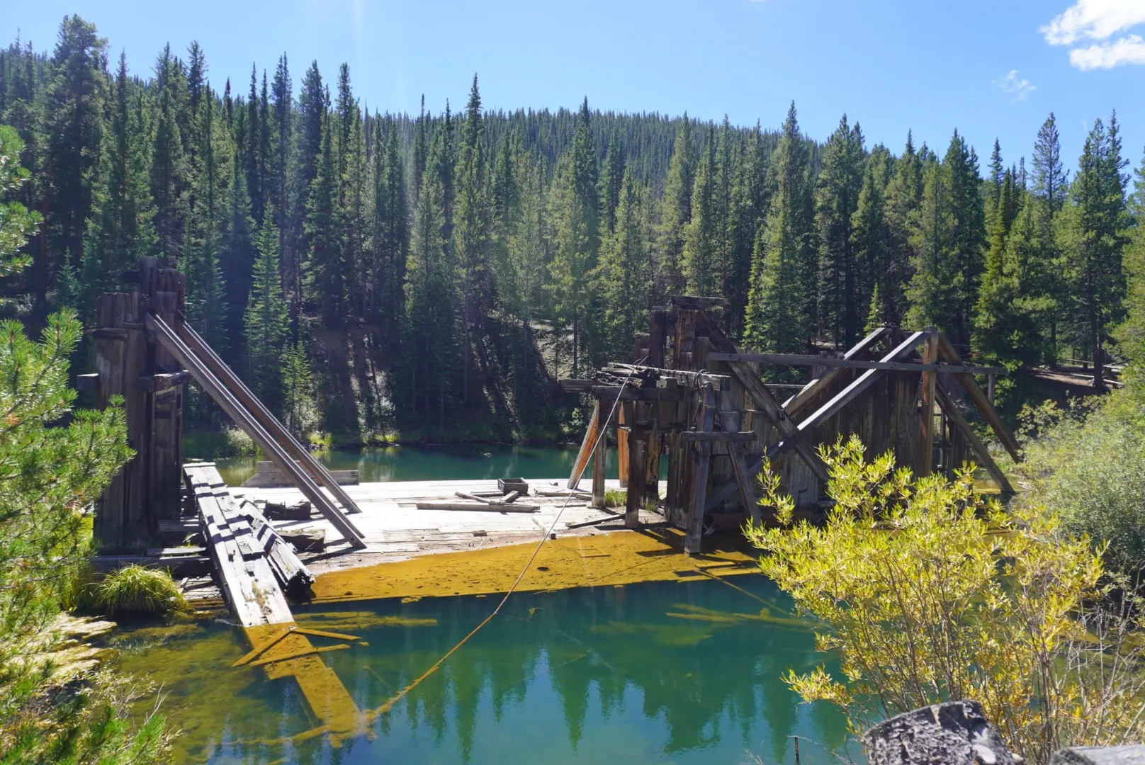 Weathered wooden mine structures beside a green pond, surrounded by pines and yellowing shrubs.